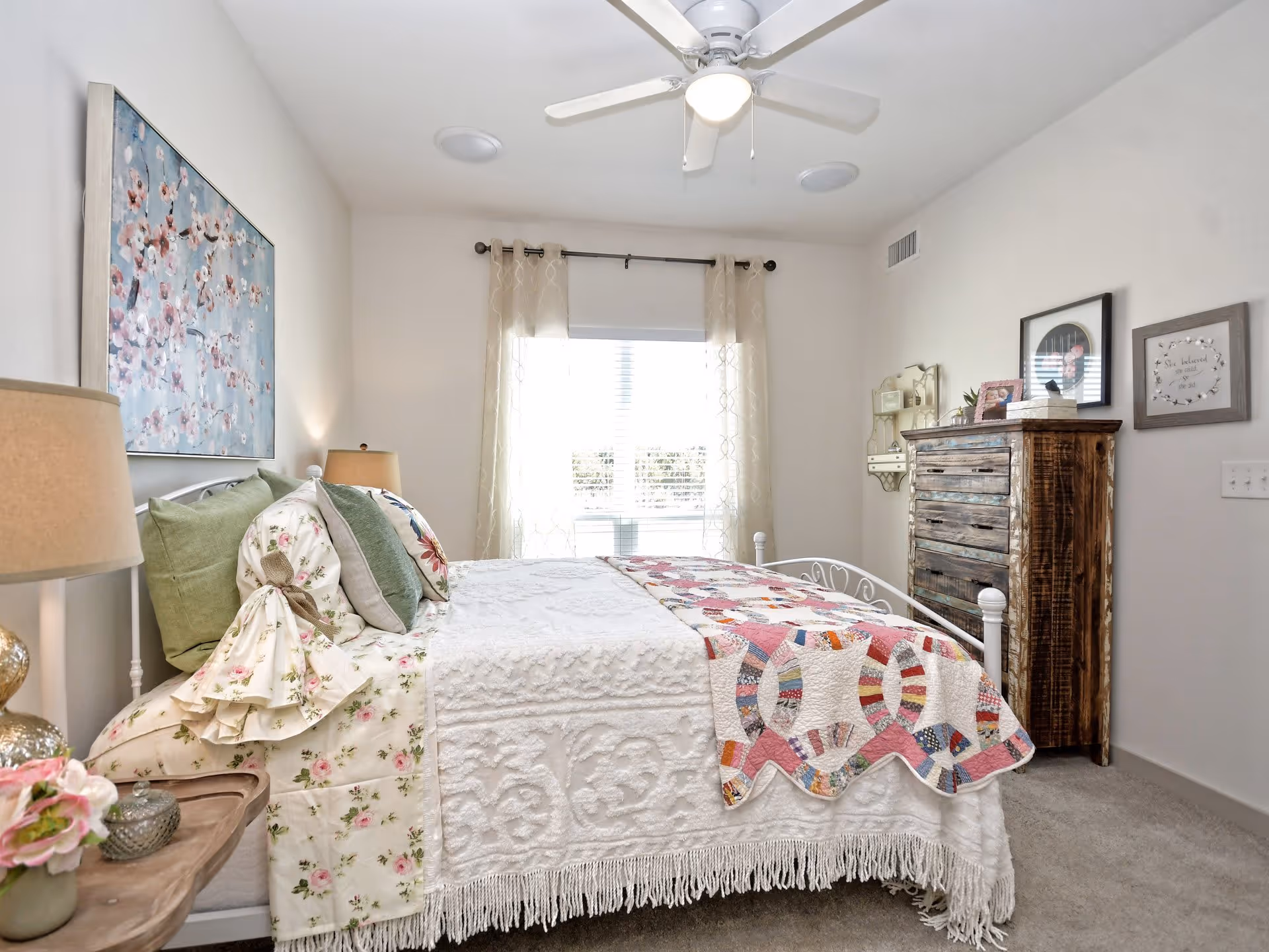 A bright and cozy bedroom with a white metal bed frame, floral and green pillows, a white textured bedspread, and a colorful quilt at the foot of the bed. There is a wooden nightstand with a lamp and flower vase on the left side, a large window with sheer curtains letting in natural light, and a rustic wooden dresser with framed pictures and decor on the right wall. A ceiling fan is mounted on the white ceiling.