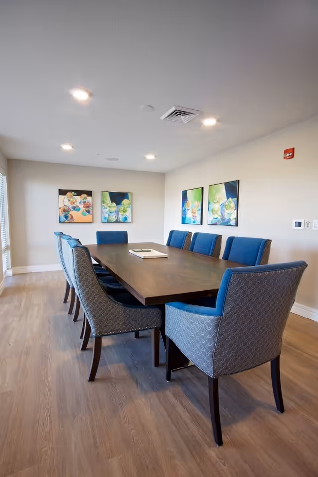 Long wooden dining table surrounded by upholstered blue and gray chairs in a bright meeting/dining room with colorful artwork on the walls.