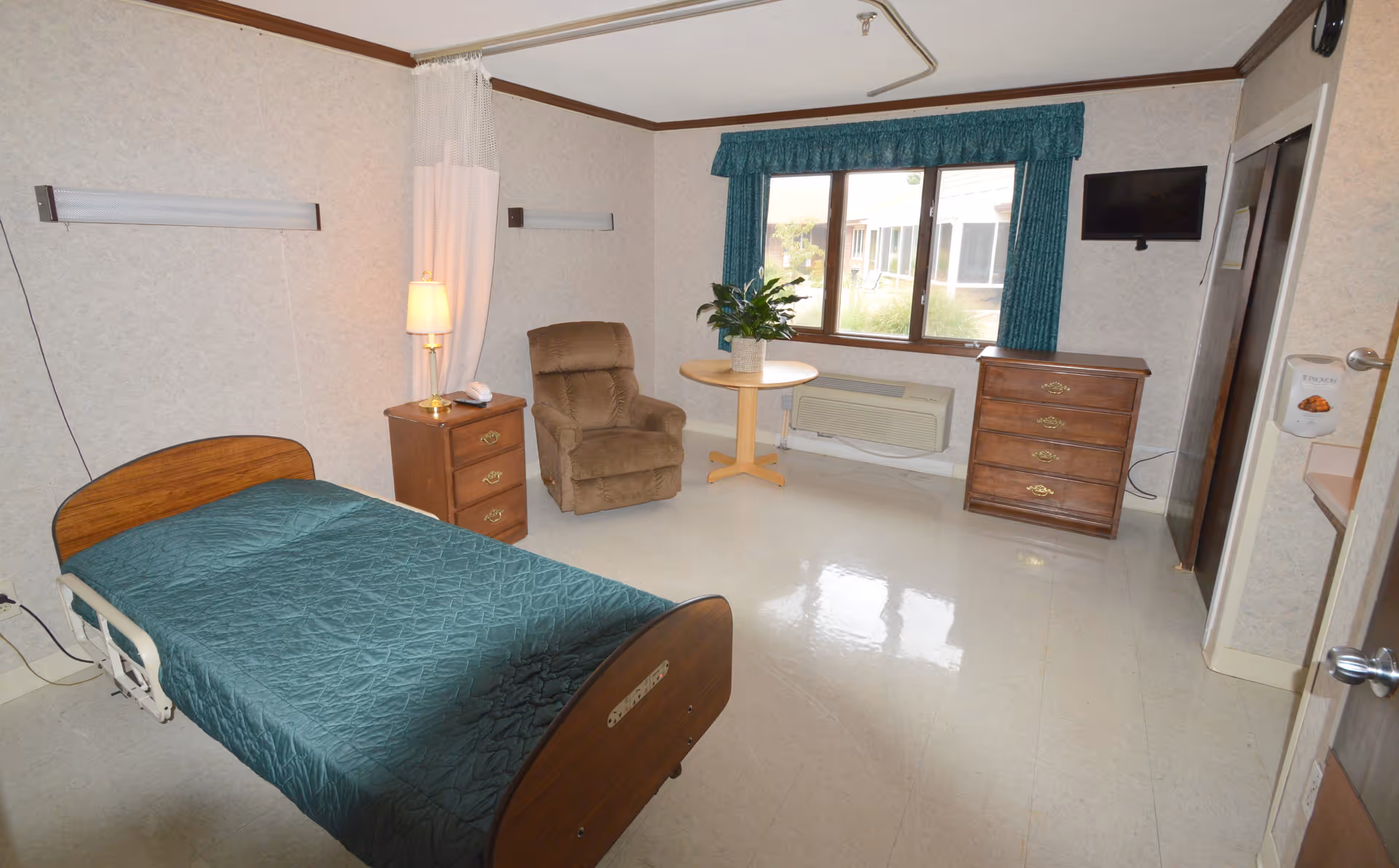 A single-occupancy senior care bedroom with a bed, recliner, nightstand, dresser, small table by a window and wall-mounted TV.