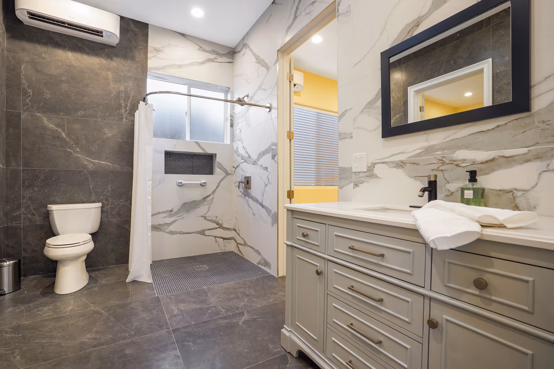 Modern senior living facility bathroom with a white toilet, walk-in shower with a curtain, gray marble floor and wall tiles, a gray vanity with a white countertop, a black framed mirror, and a window providing natural light.