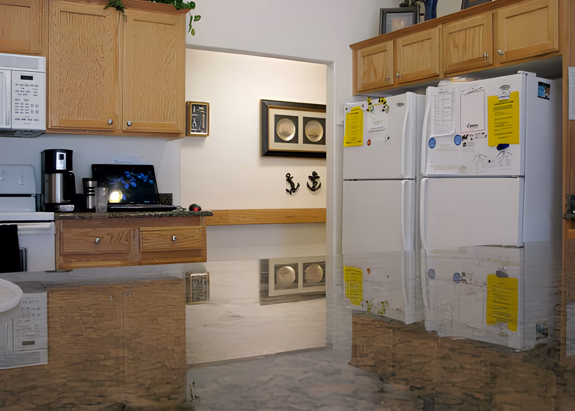 Interior view of a kitchen featuring wooden cabinets, a microwave, a coffee maker, two white refrigerators with papers and notes attached, and a polished countertop reflecting the surroundings. In the background, there is a wall with framed artwork and decorative anchors.