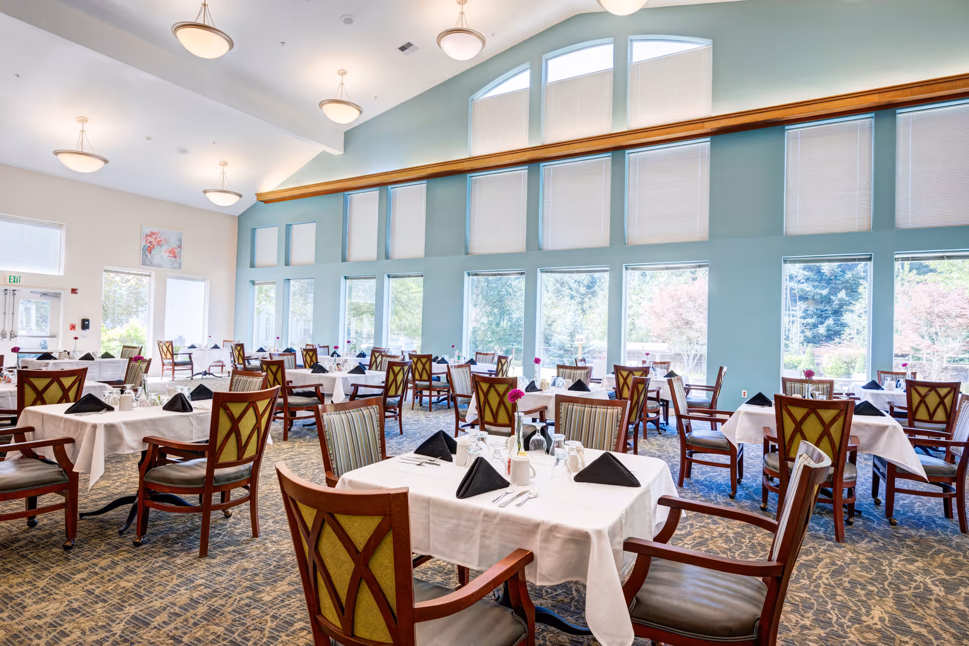 Bright dining room with multiple tables covered in white tablecloths, each set with black napkins, glassware, and silverware. The room features large windows with views of trees outside, high ceilings with hanging light fixtures, and patterned carpet flooring.