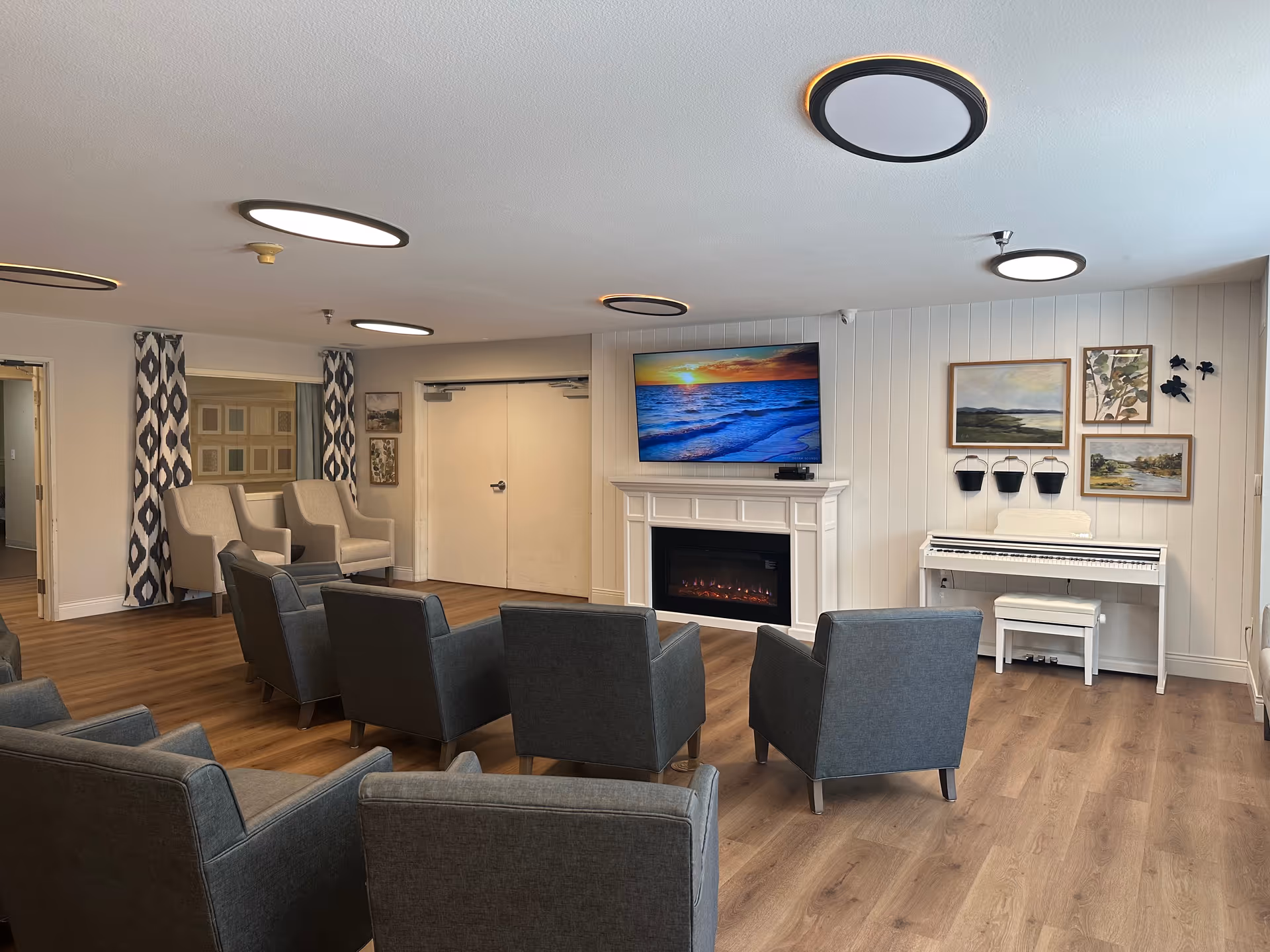 A senior living facility common area with multiple gray armchairs arranged in rows facing a white electric fireplace and a wall-mounted TV displaying a sunset over the ocean. To the right of the fireplace is a white piano with a matching bench, and above it are framed paintings and decorative wall hangings. The room has wood flooring, white paneled walls, and modern circular ceiling lights.
