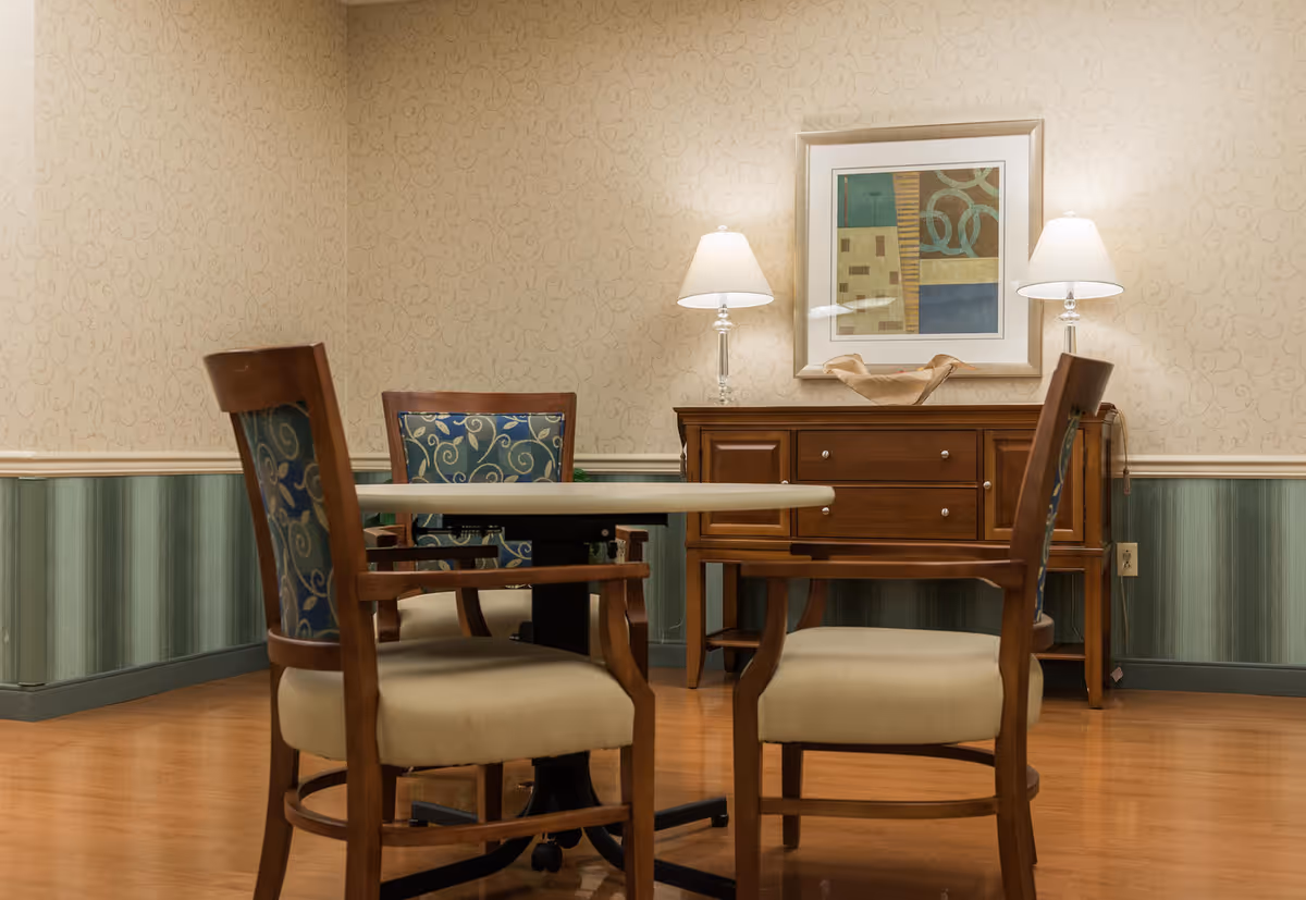 A small seating area with a round table and padded wooden chairs in front of a wooden sideboard topped with two lamps and framed artwork.