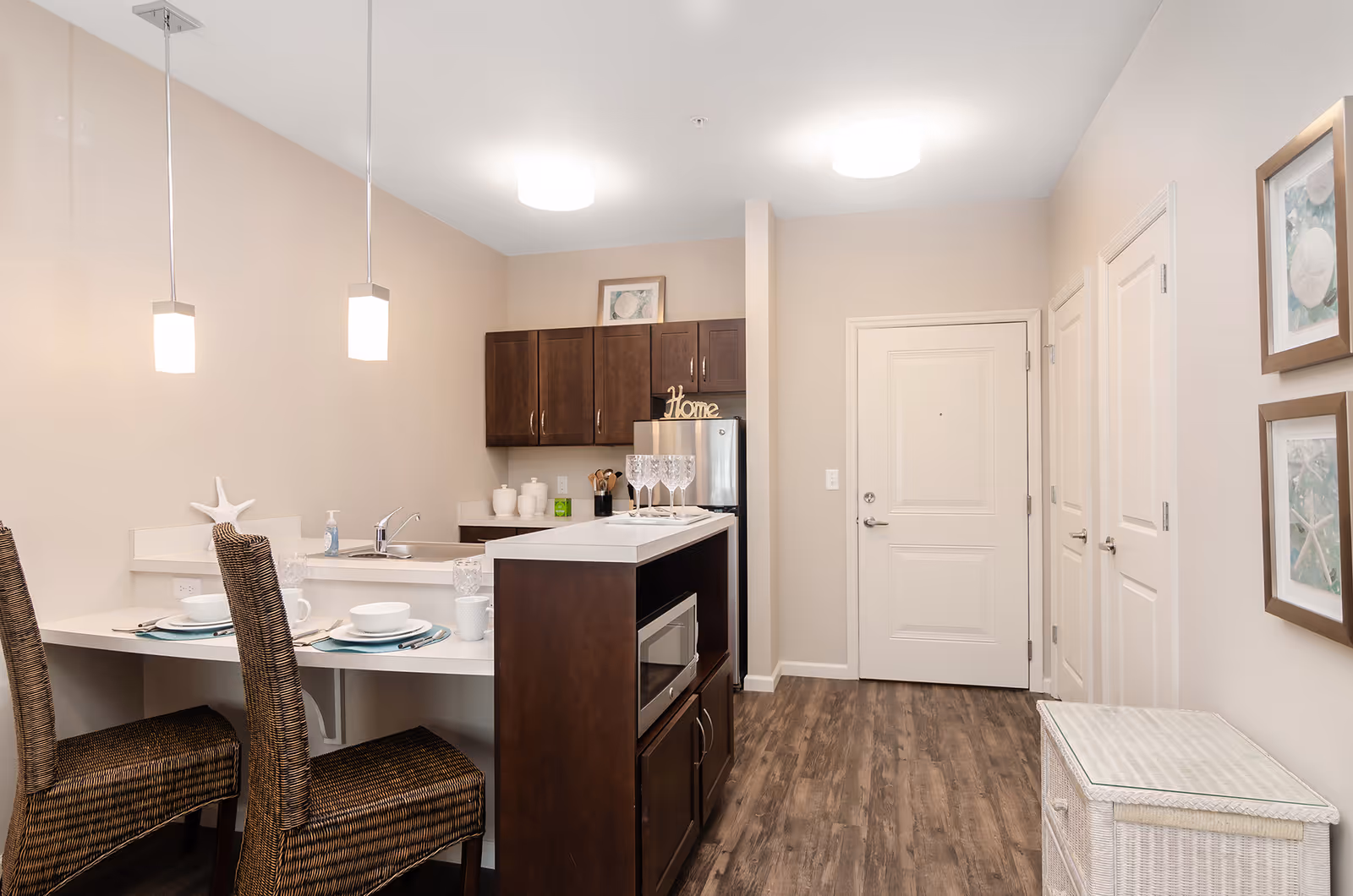Interior view of a modern kitchen and dining area in a senior living facility. The kitchen features dark wood cabinets, a small refrigerator, a microwave, and a white countertop with a sink. Two wicker chairs are placed at a breakfast bar set with white dishes and glassware. The room has light beige walls, wood flooring, two ceiling lights, and framed artwork on the wall near the entrance door.