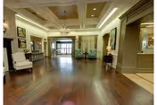 Spacious interior common area with polished dark hardwood floors, white armchairs, chandeliers, and large windows letting in natural light. The room features decorative columns, a fireplace, and a seating area with blue chairs near the windows.