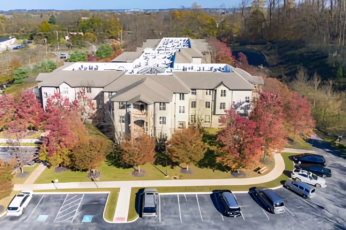 Aerial view of a multi-story senior living facility named Spring Mill surrounded by trees with autumn foliage. The building has a beige exterior and a large parking lot with several vehicles parked. There are sidewalks and landscaped areas with green grass and colorful trees around the building.
