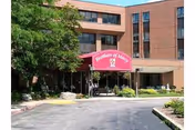 Exterior view of a multi-story brick building with a covered entrance. A red sign above the entrance reads 'Brothers of Mercy'. There are trees and shrubs around the entrance area and a paved driveway leading up to it.