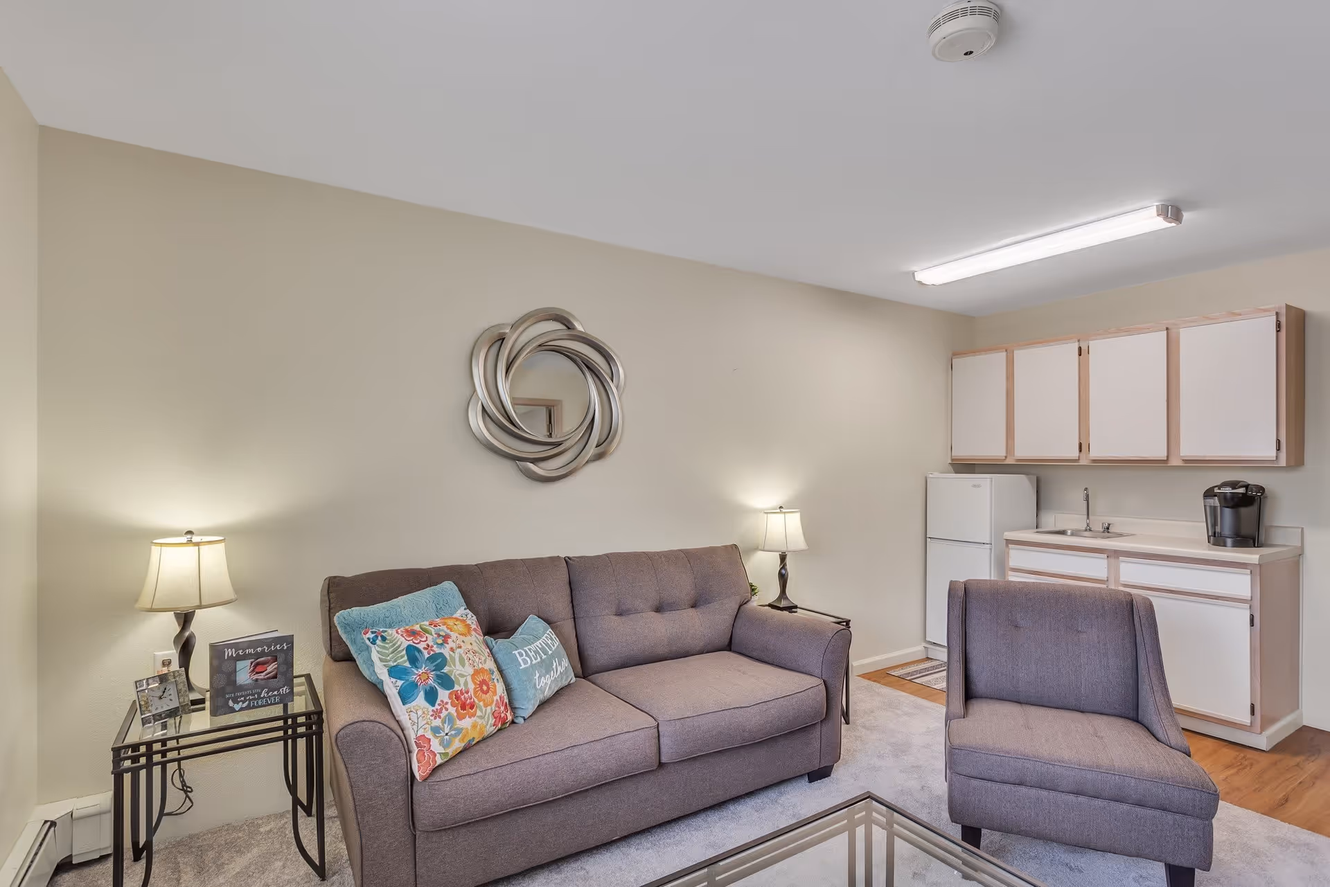 A cozy living room area with a gray sofa adorned with colorful and patterned throw pillows, a matching gray armchair, and a glass coffee table. There are two side tables with lamps on either side of the sofa. On the wall above the sofa is a decorative circular mirror. In the background, there is a small kitchenette with white cabinets, a sink, a refrigerator, and a coffee maker. The room has beige walls and a mix of carpet and wood flooring.