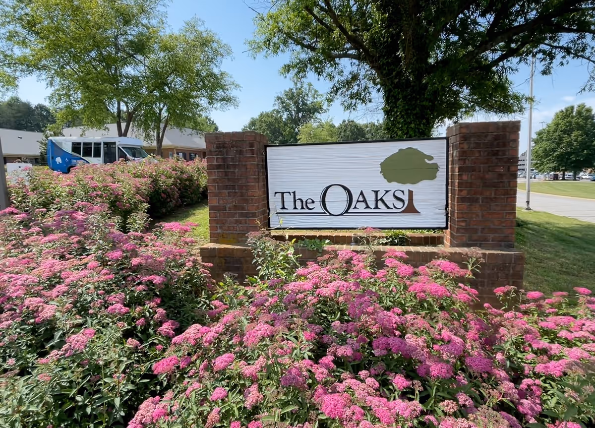 A brick sign for The Oaks surrounded by lush pink flowering bushes and green trees under a clear blue sky. A white and blue vehicle is partially visible in the background near a building.