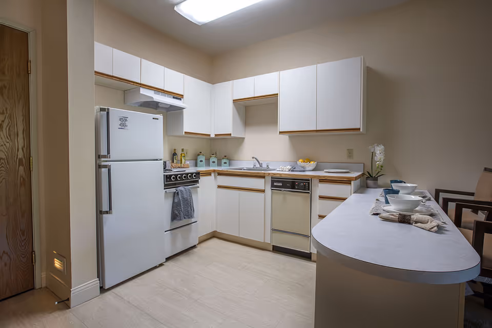 Compact U-shaped kitchen with white cabinets, refrigerator, stove, dishwasher and a breakfast bar set with bowls.