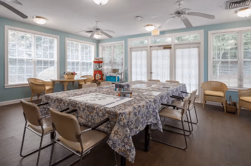 A bright activity room with large windows letting in natural light. There are two tables covered with patterned tablecloths, set up for a puzzle activity with puzzle pieces and boxes on top. Surrounding the tables are several chairs with armrests. In the background, there is a small round table with wicker chairs and a popcorn machine near the windows. The walls are painted light blue, and ceiling fans with lights are mounted on the ceiling.