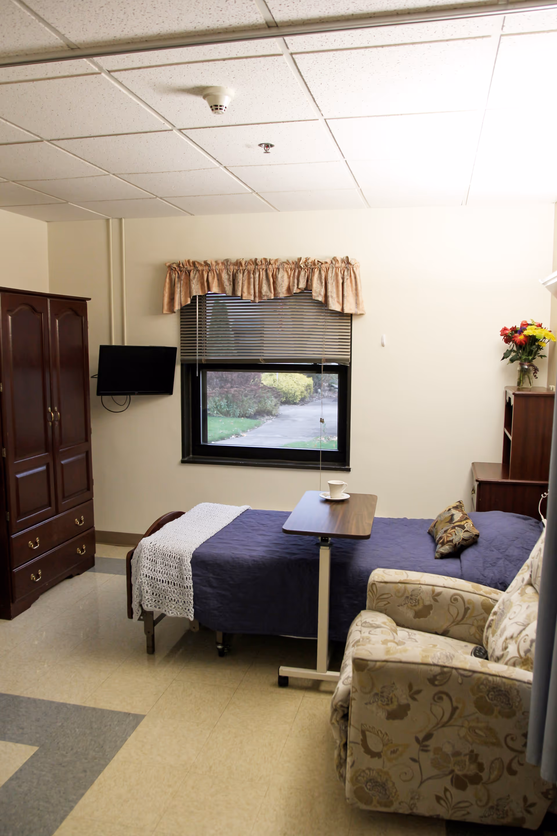 A senior living facility bedroom with a single bed, overbed table, armchair, wardrobe, and a window looking outside.