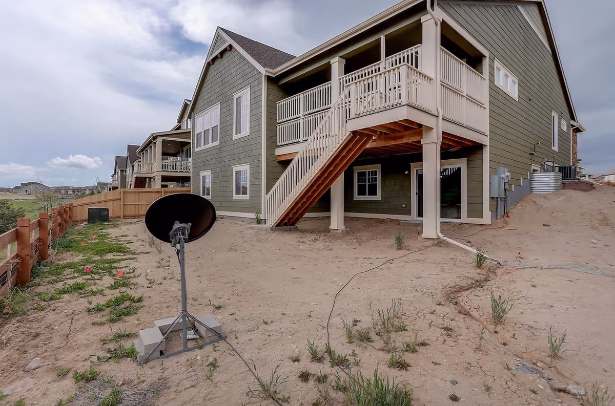 Exterior view of a green two-story residential building with a wooden staircase leading to a balcony. The ground is mostly bare dirt with some sparse grass and a satellite dish mounted on a tripod. A wooden fence runs along the left side of the property under a partly cloudy sky.