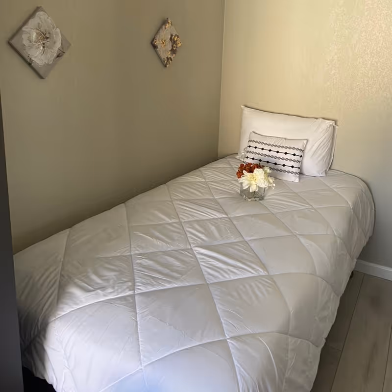 A small bedroom with a single bed covered in a white quilted comforter. The bed has two pillows, one plain white and one decorative with a black and white pattern. A small glass vase with white and orange flowers is placed on the bed. Two small square floral wall decorations hang on the beige wall above the bed.