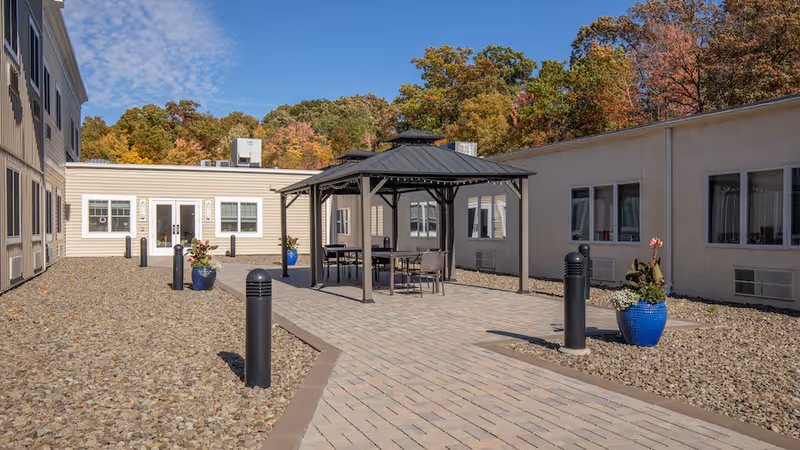 Outdoor courtyard area at Linwood Nursing and Rehabilitation Center featuring a paved walkway leading to a gazebo with tables and chairs. The courtyard is surrounded by single-story building walls with windows, decorative planters with flowers, and trees with autumn foliage in the background under a clear blue sky.