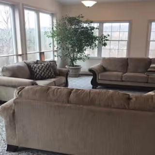 A bright sitting room with three beige sofas, large windows, and a potted plant.