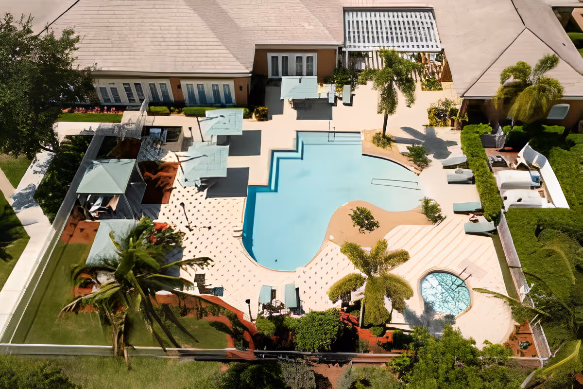 Aerial view of a senior living facility's outdoor pool area featuring a large swimming pool with steps, a smaller circular hot tub, several lounge chairs, palm trees, and surrounding greenery. The pool area is adjacent to a building with multiple windows and a covered patio.