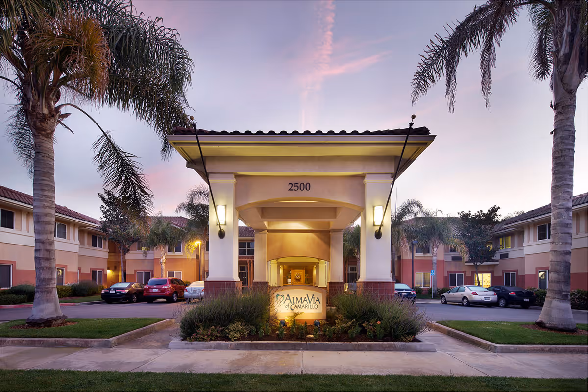 Front entrance of the AlmaVia of Camarillo senior living facility with a porte-cochère, landscaping, palm trees, and parked cars at dusk.