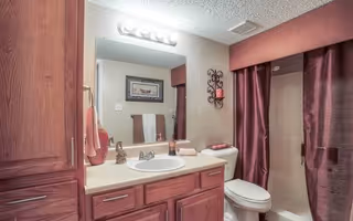 Bathroom with wooden cabinets, a white sink with silver faucet, a large mirror above the sink, a toilet next to the sink, and a shower with a burgundy curtain. There are towels hanging on the wall and decorative items including a framed picture and a wall-mounted candle holder.