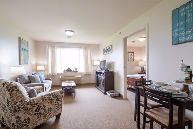 A bright living room with a large window covered by light curtains. The room features a patterned armchair, a gray sofa with cushions, a wooden coffee table with a chessboard, and a TV on a dark wooden stand. To the right, there is a small dining table set for two with plates and glasses. An open doorway leads to a bedroom with a bed and bedside lamp visible.