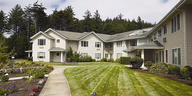 Two-story beige assisted living building front with a curved walkway, manicured lawn, and landscaped shrubs.