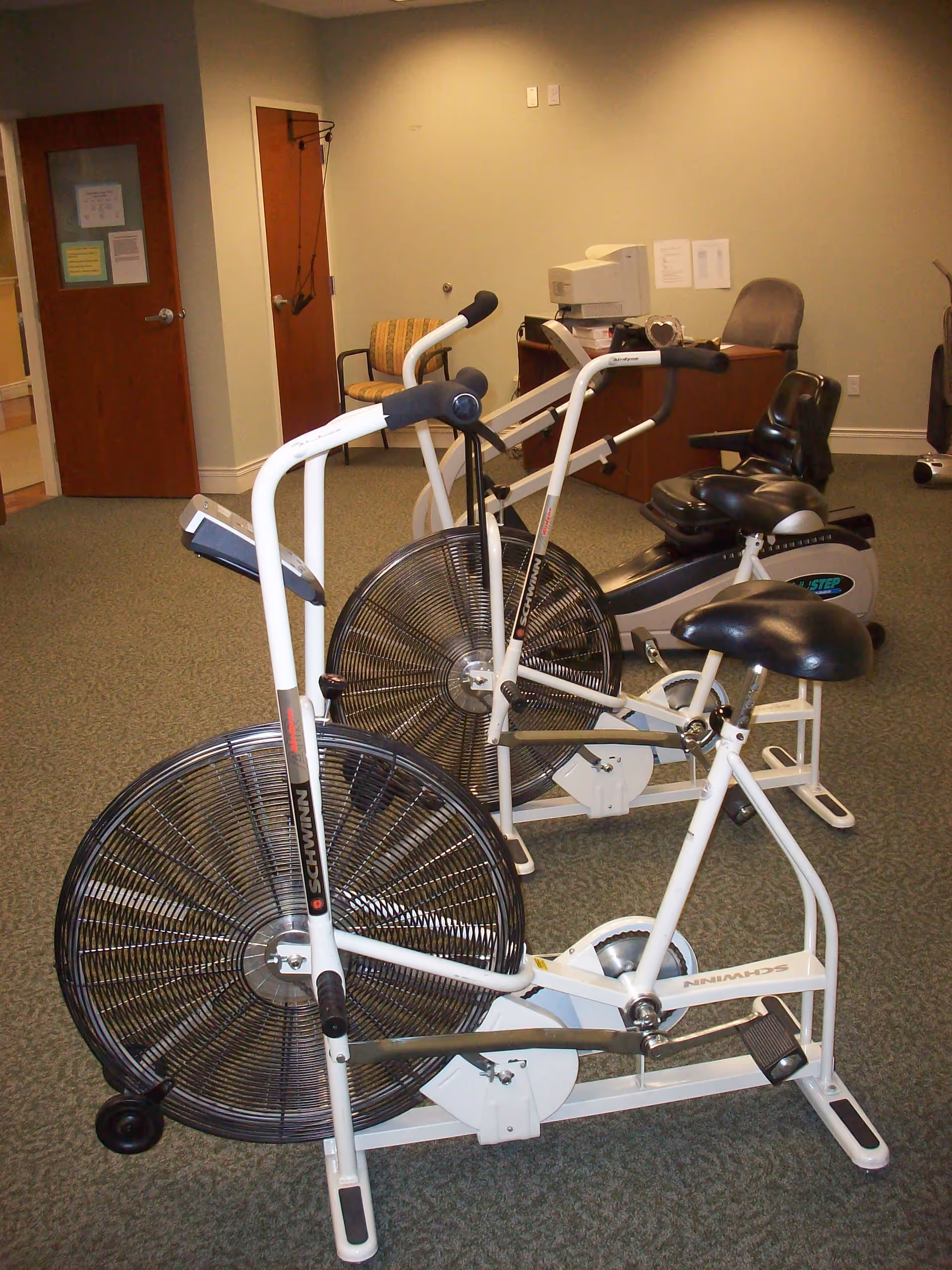 Indoor exercise room with two stationary exercise bikes, a recumbent bike, a chair, and a desk with a computer monitor in the background.