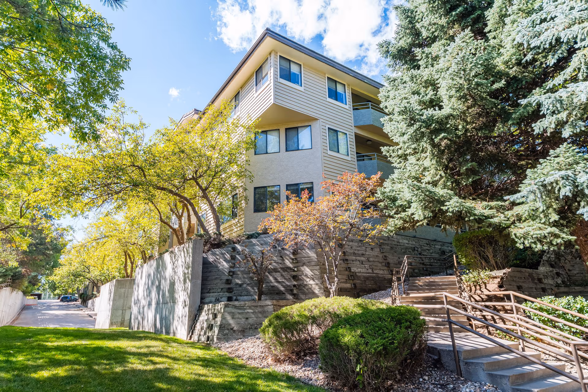Exterior view of a multi-story residential building surrounded by trees and greenery under a blue sky with some clouds. There are stairs with railings leading up to the building, and a well-maintained lawn and bushes in the foreground.
