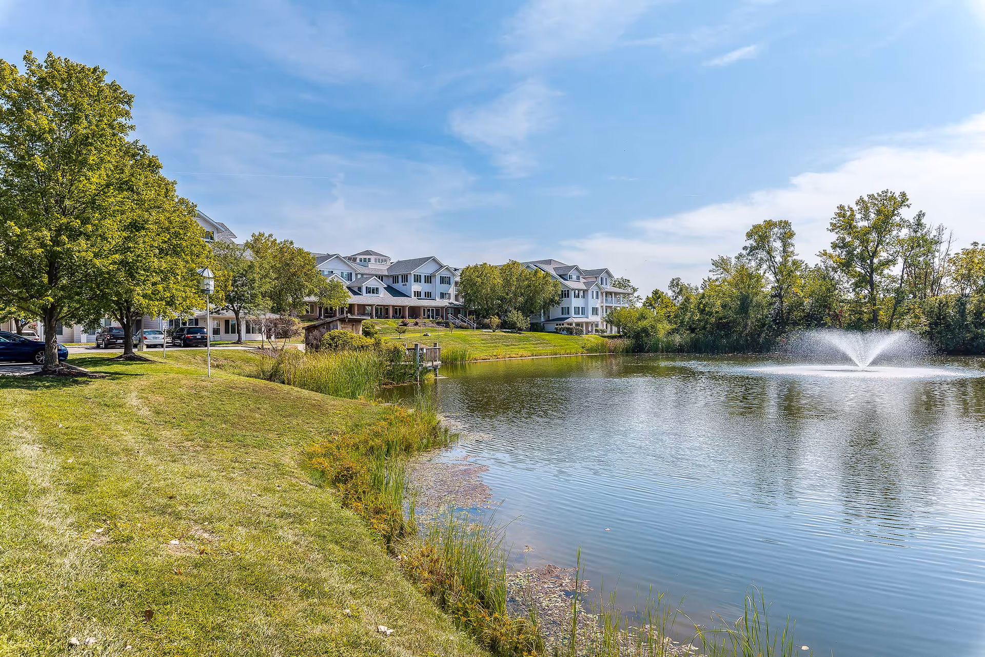 A senior living facility building situated beside a pond with a water fountain. The area is surrounded by green grass, trees, and a clear blue sky.