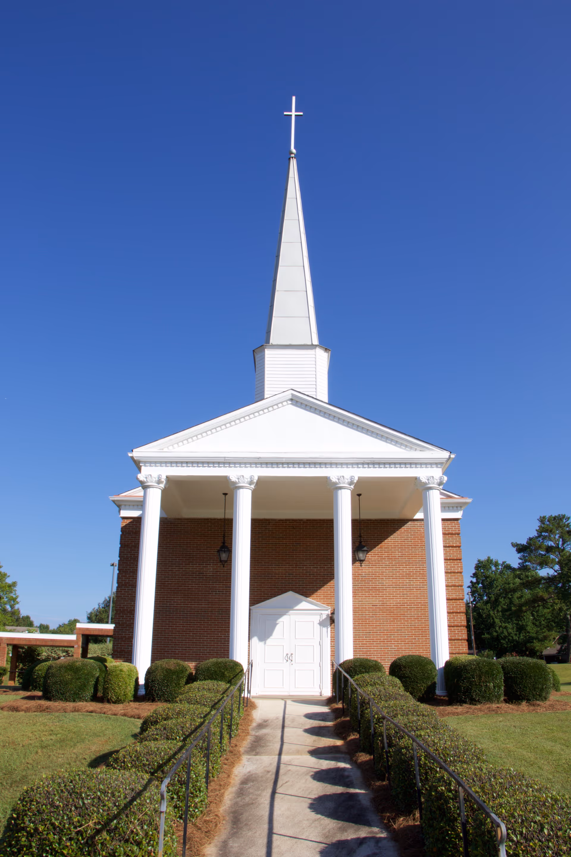 Front view of a red-brick church-like building with white columns, a tall white steeple topped by a cross, and a walkway lined with trimmed bushes.