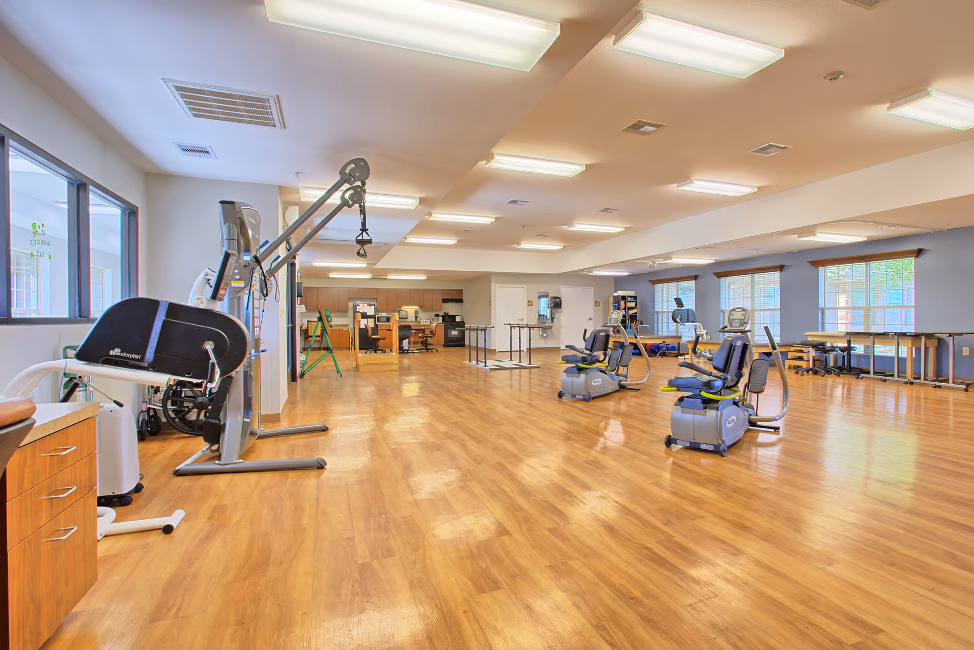 A spacious rehabilitation or physical therapy room with wooden flooring, exercise equipment including recumbent bikes and a multi-functional exercise machine, large windows letting in natural light, and a desk area in the background.