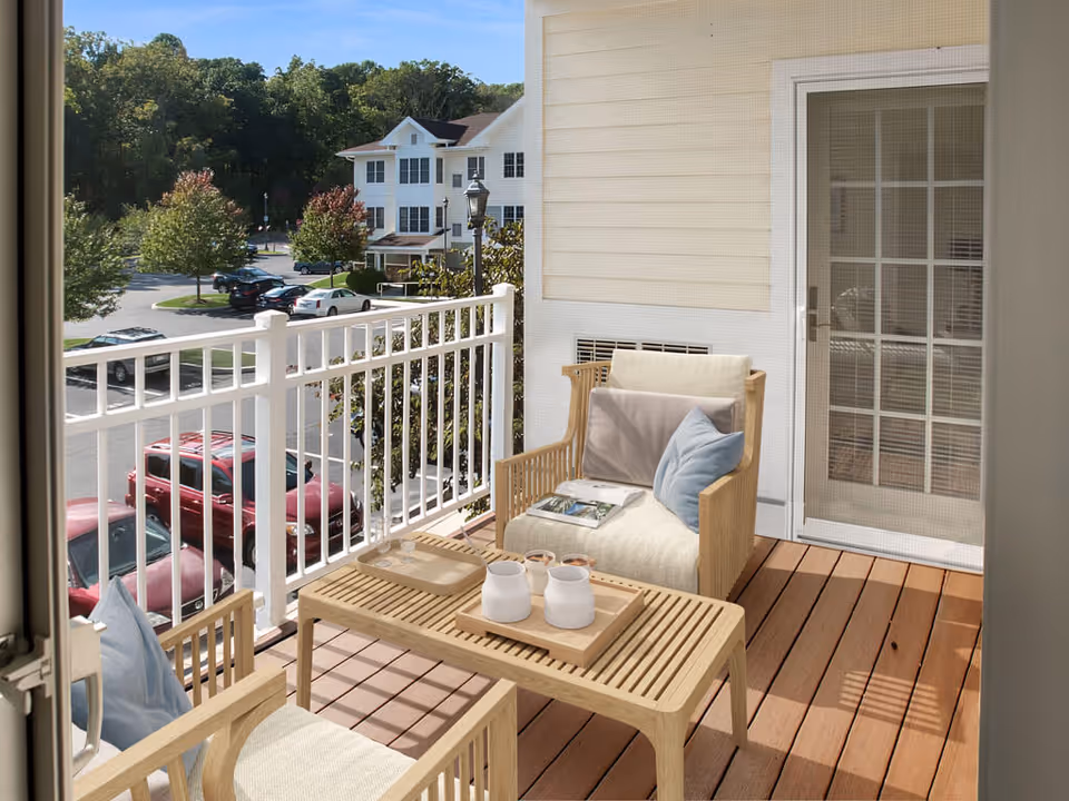 A small balcony with two wooden chairs featuring cushions and a wooden table between them. On the table are two white mugs and a tray. The balcony has a white railing and overlooks a parking lot with cars and trees. A glass door with a grid pattern leads inside the building.