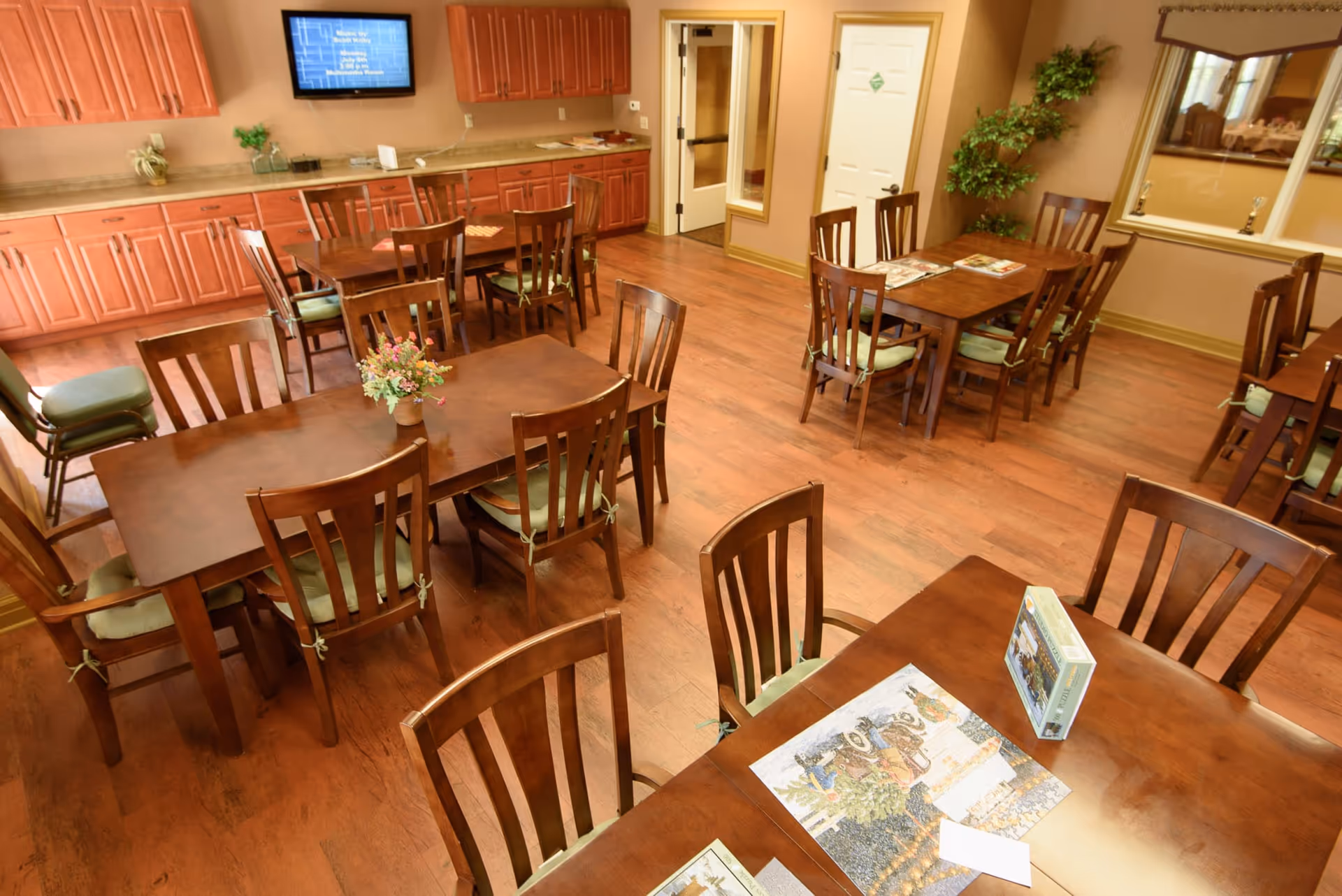 A well-lit dining room with multiple wooden tables and chairs with green cushions. Some tables have puzzles and magazines on them. The room has wooden flooring, a wall-mounted TV, wooden cabinets along one wall, a door, and a large window with a plant nearby.