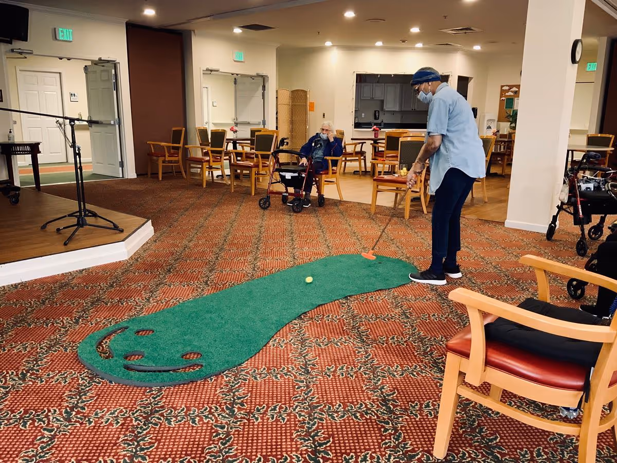 An indoor common area with patterned carpet and wooden chairs. A person wearing a mask and a blue shirt is playing mini golf on a small green putting mat with a smiley face design. Another elderly person with a walker is seated and watching. The room has exit signs and doors in the background.