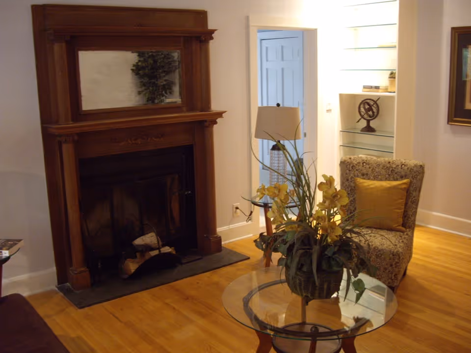 A cozy living room with a wooden fireplace featuring a large mirror above it, a patterned armchair with a yellow cushion, a glass coffee table with a floral arrangement, a side table with a lamp, and built-in shelves with decorative items.