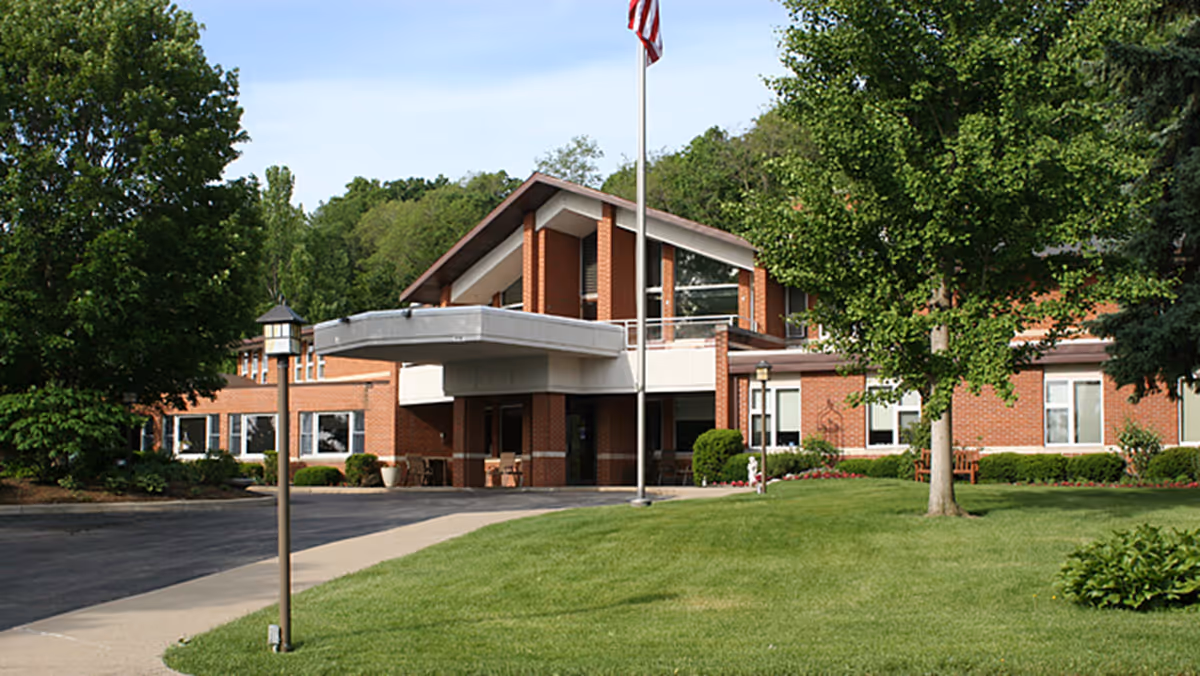 Front exterior view of a brick building with a covered entrance, surrounded by green trees and a well-maintained lawn. An American flag is flying on a flagpole in front of the building.