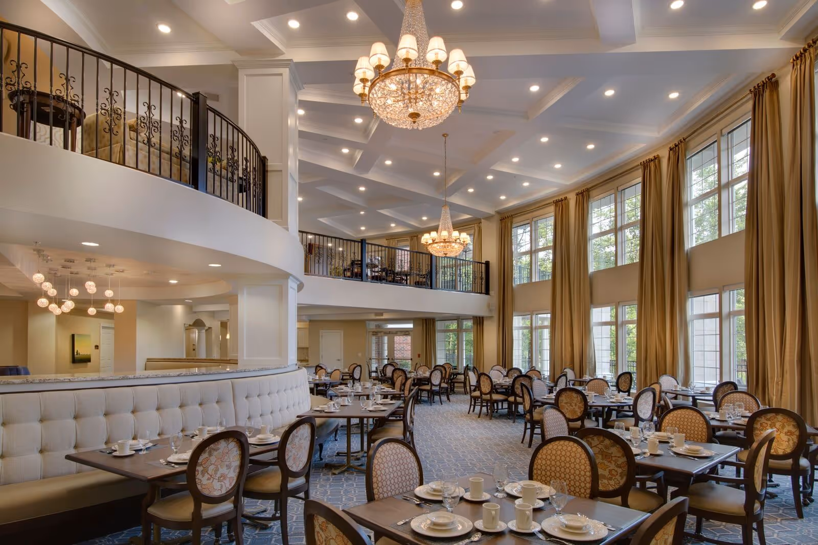 Spacious and elegant dining room with multiple tables set with plates, cups, and glasses. The room features large windows with long beige curtains, high ceilings with chandeliers, and a mezzanine level with additional seating. The decor includes upholstered chairs and a curved banquette seating area.