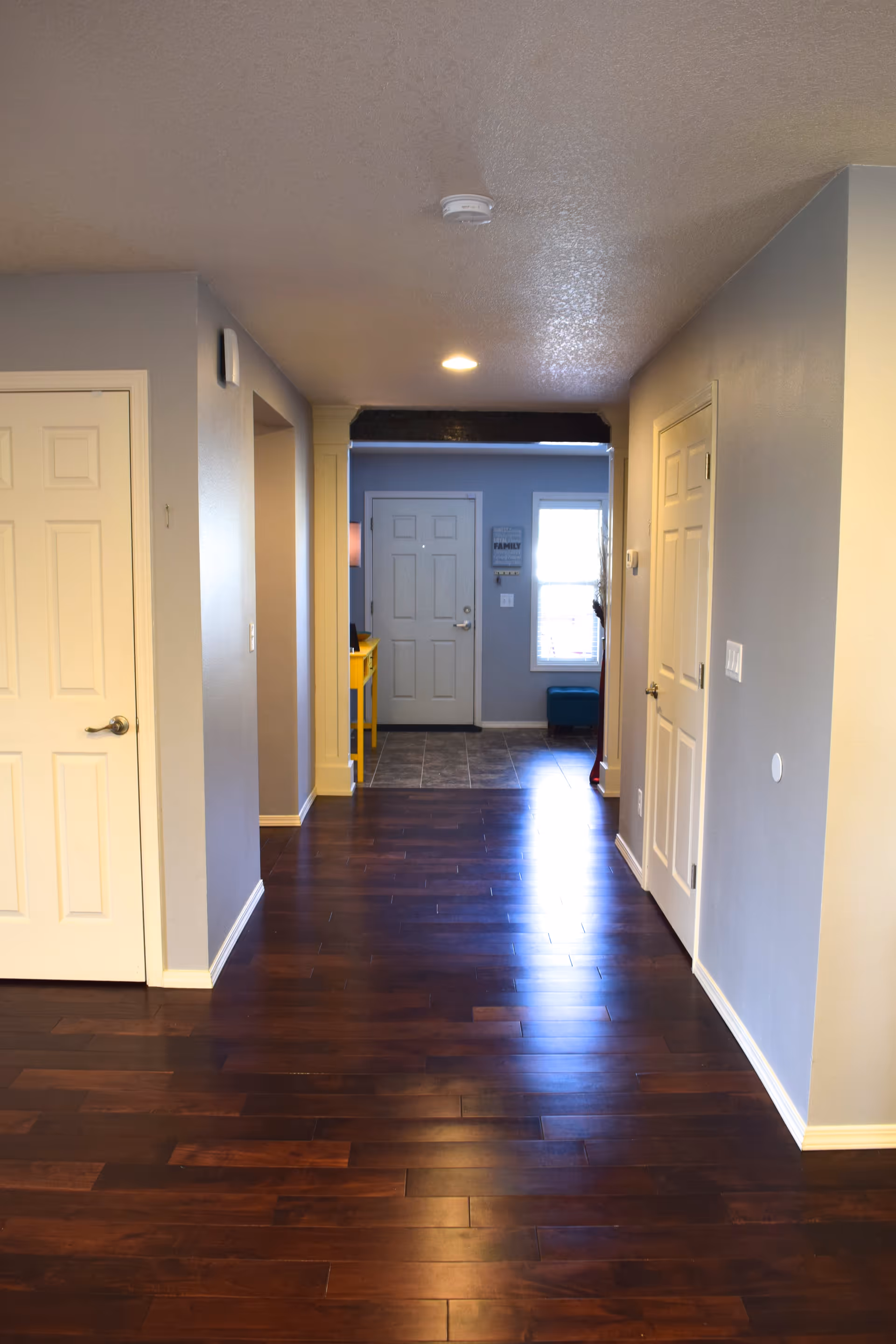 Interior hallway with dark wooden flooring, light gray walls, and white doors on both sides. At the end of the hallway is a front door with a window to its right and a small yellow table against the left wall.