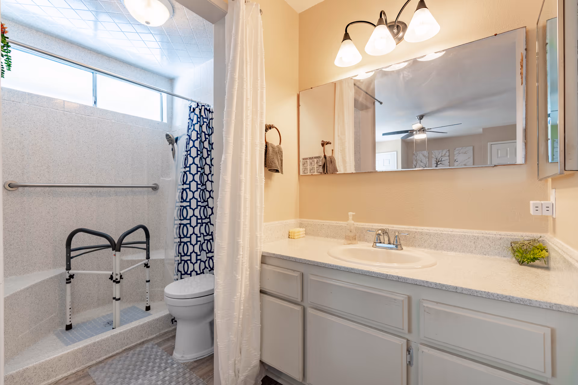 A bathroom with a walk-in shower featuring a blue and white patterned shower curtain, a grab bar, and a shower chair. There is a toilet next to the shower. The vanity has a white countertop with a sink, soap dispenser, and a small plant. Above the vanity is a large mirror with three light fixtures. The walls are painted beige.