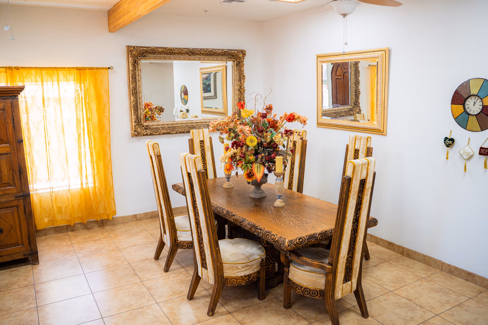 A dining room with a wooden table surrounded by eight high-back chairs. The table is decorated with a large floral centerpiece and two candle holders. The room has tiled flooring, white walls, two large ornate mirrors, a colorful wall clock, and yellow curtains covering a window. There is also a wooden cabinet on the left side.