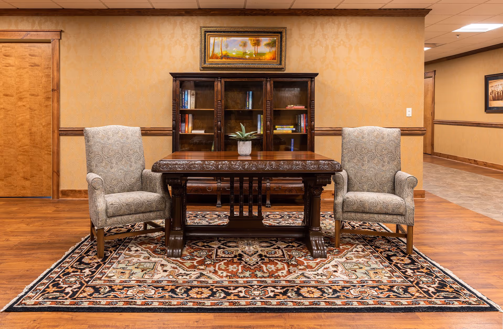 A furnished sitting area with two upholstered armchairs facing a carved wooden table on a patterned rug in front of a bookshelf.