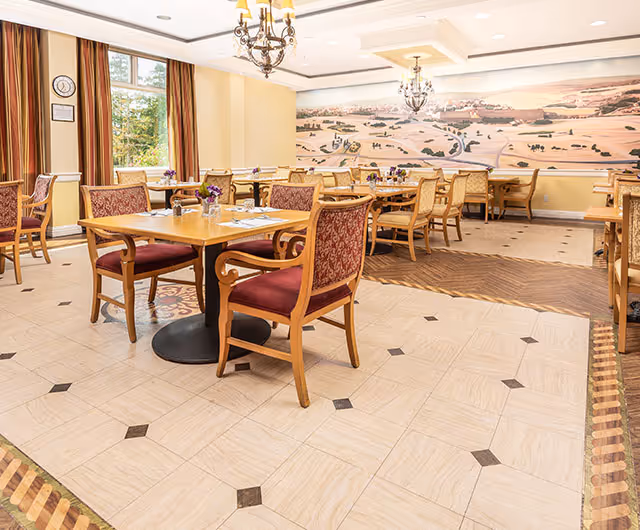 Bright dining room with wooden tables and upholstered chairs set for meals, chandeliers, and a mural on the far wall.