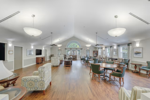 A spacious and well-lit common area in a senior living facility with wooden flooring, multiple round tables surrounded by green cushioned chairs, several armchairs, ceiling fans, and large windows allowing natural light. The room has a high vaulted ceiling with modern light fixtures and a large arched window at the far end.