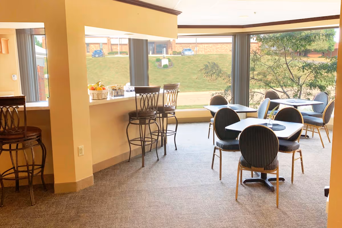 A bright dining area with several tables and chairs arranged near large windows that offer a view of a grassy outdoor area with trees and parked cars. There is a counter with bar stools on the left side, and a basket of fruit and some water bottles are placed on the counter.