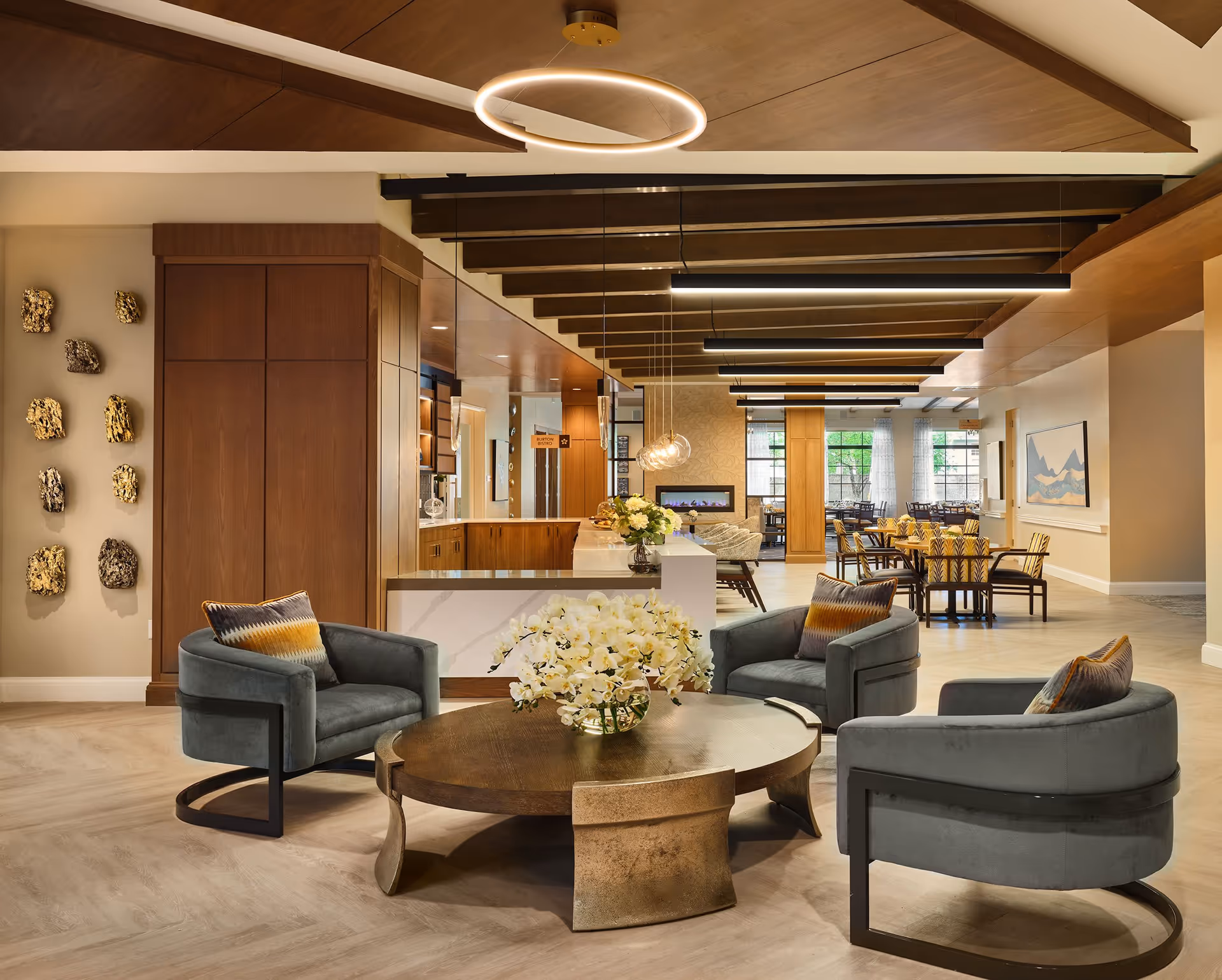 Bright modern communal lounge with a round coffee table, four upholstered chairs, floral centerpiece, and a dining area visible in the background.