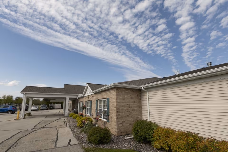 Front exterior of a single-story assisted living building with a covered entrance, parking area, and a partly cloudy sky.