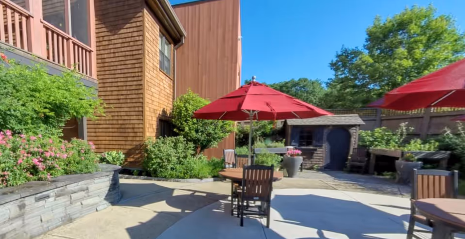 Sunny outdoor courtyard with round tables, chairs and red umbrellas beside landscaped beds and a wood-clad building.