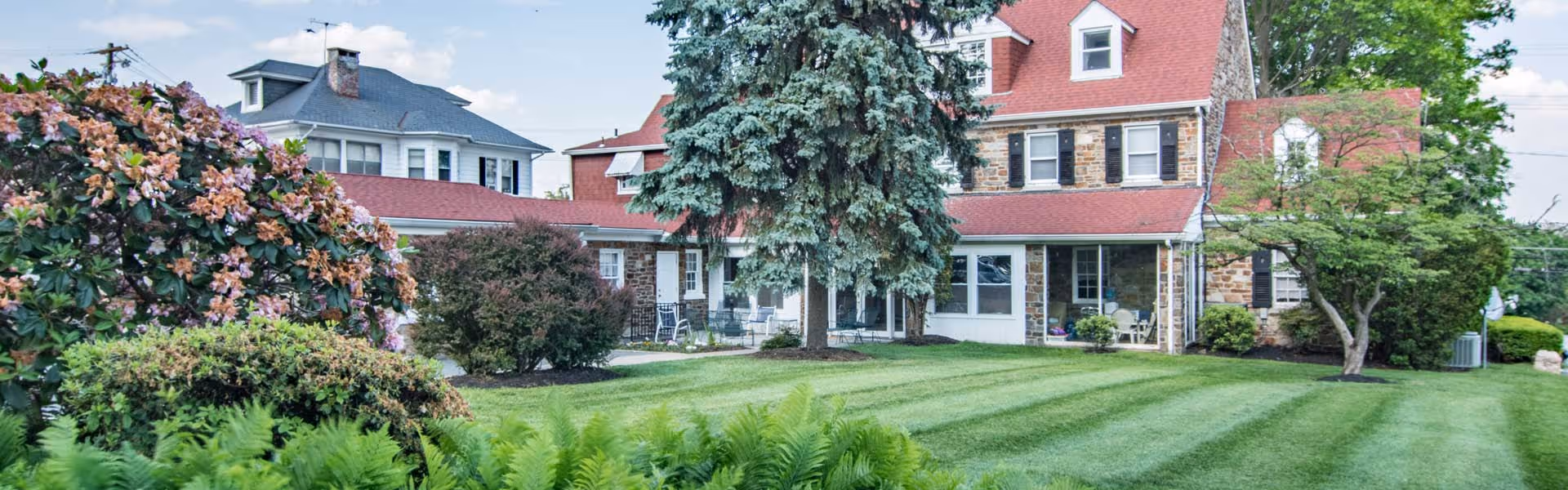 A large, well-maintained lawn with striped mowing patterns in front of a multi-story stone and brick building with a red roof. The building has multiple windows and a porch area with outdoor seating. There are various trees and bushes surrounding the lawn, including a flowering bush on the left and a tall evergreen tree near the center.