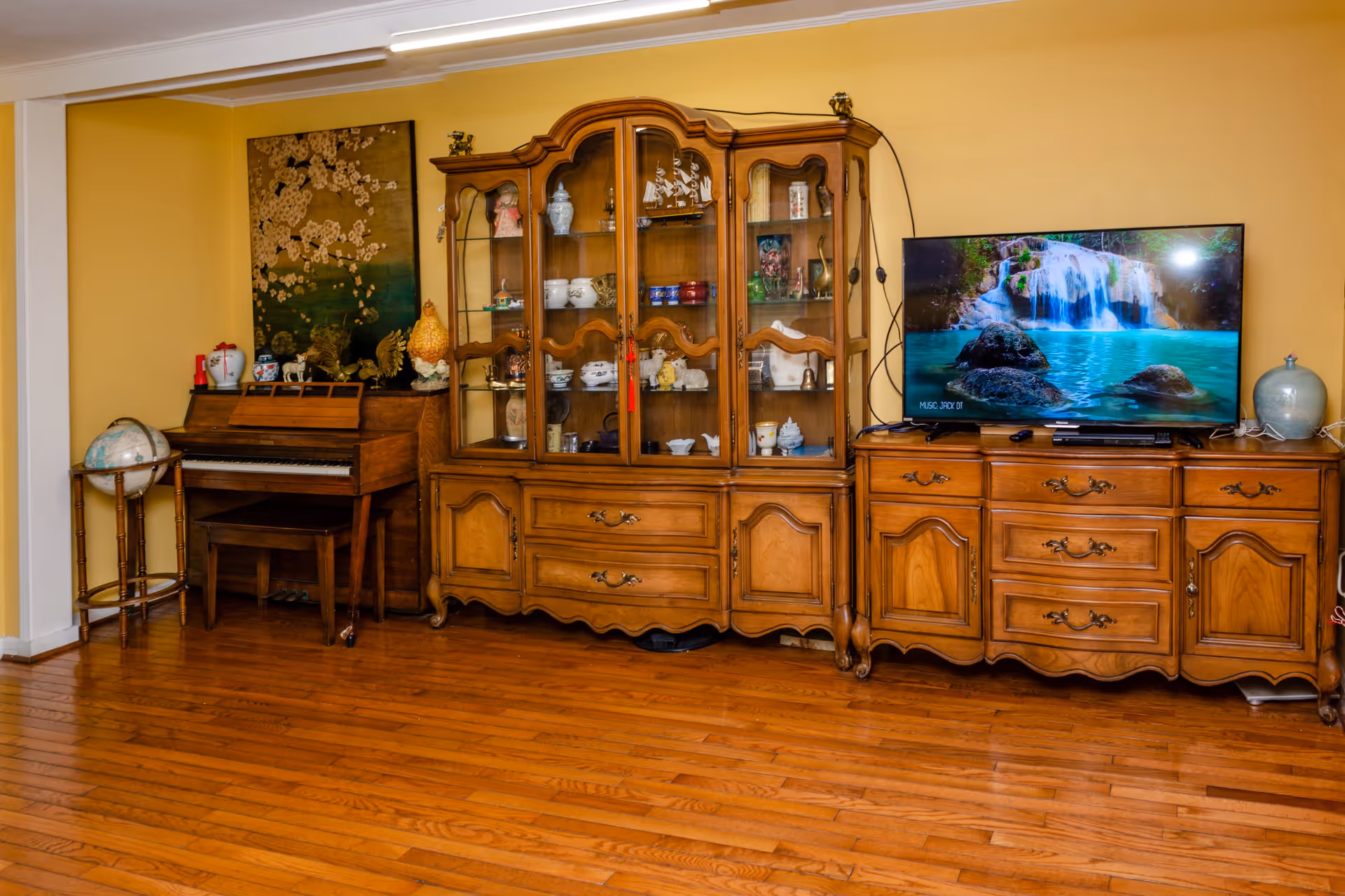 A cozy living room area with wooden flooring and yellow walls featuring a wooden piano with a bench, a globe on a stand, a large wooden display cabinet filled with decorative items, and a wooden TV stand with a flat-screen TV showing a waterfall scene.