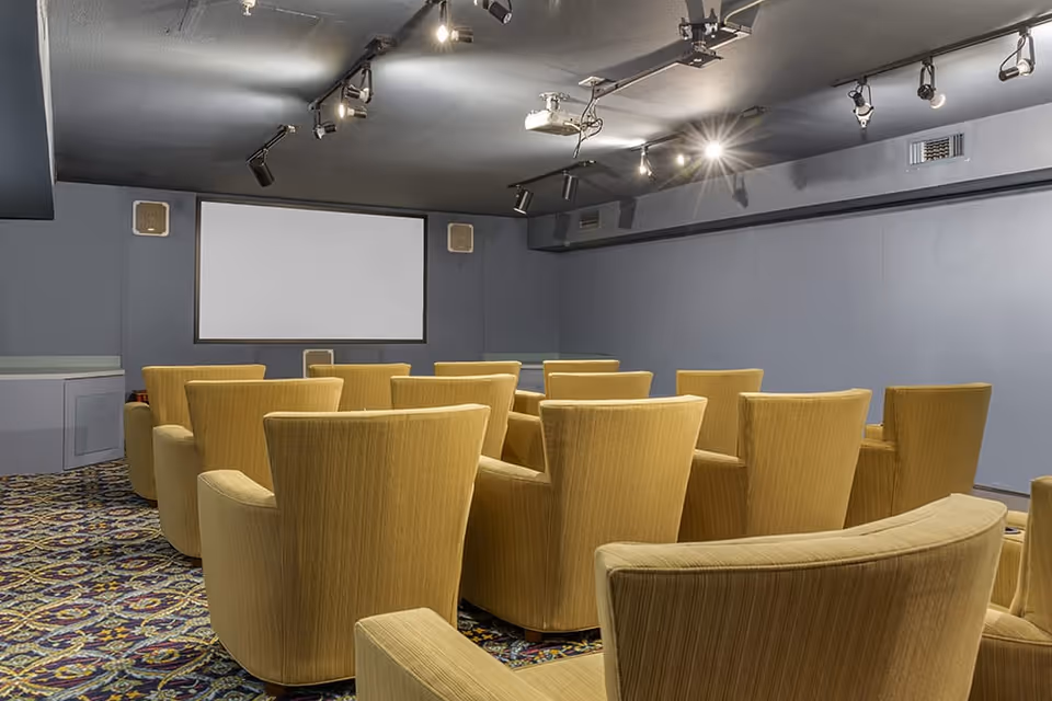 A small theater room with multiple rows of comfortable yellow armchairs facing a large blank projection screen on a gray wall. The ceiling has several spotlights and a projector mounted.
